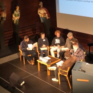 Schüler sitzen auf einer Bühne auf Stühlen um einen Tisch herum und nehmen an einer Podiumsdiskussion teil.