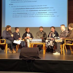 Schüler sitzen auf einer Bühne auf Stühlen um einen Tisch herum und nehmen an einer Podiumsdiskussion teil.